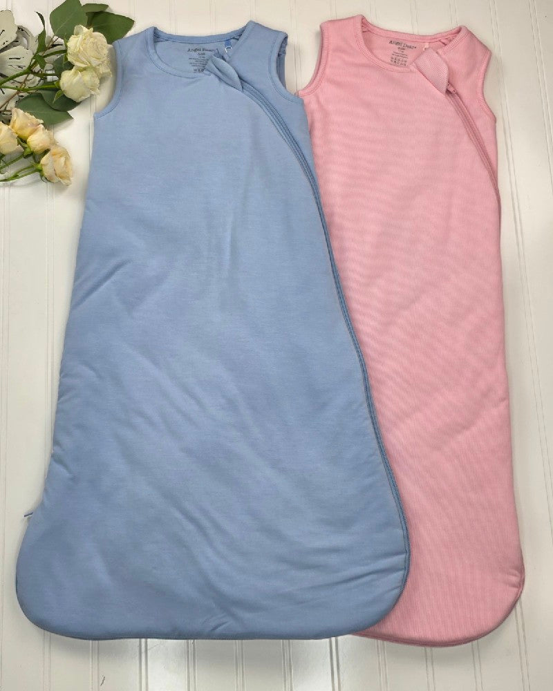 The Angel Dear Solid Blue Fog Sleep Bag and a light pink sleeveless bamboo sleep sack rest side by side on a white wooden surface, with pale yellow roses nearby for a calm, breathable baby sleep atmosphere.