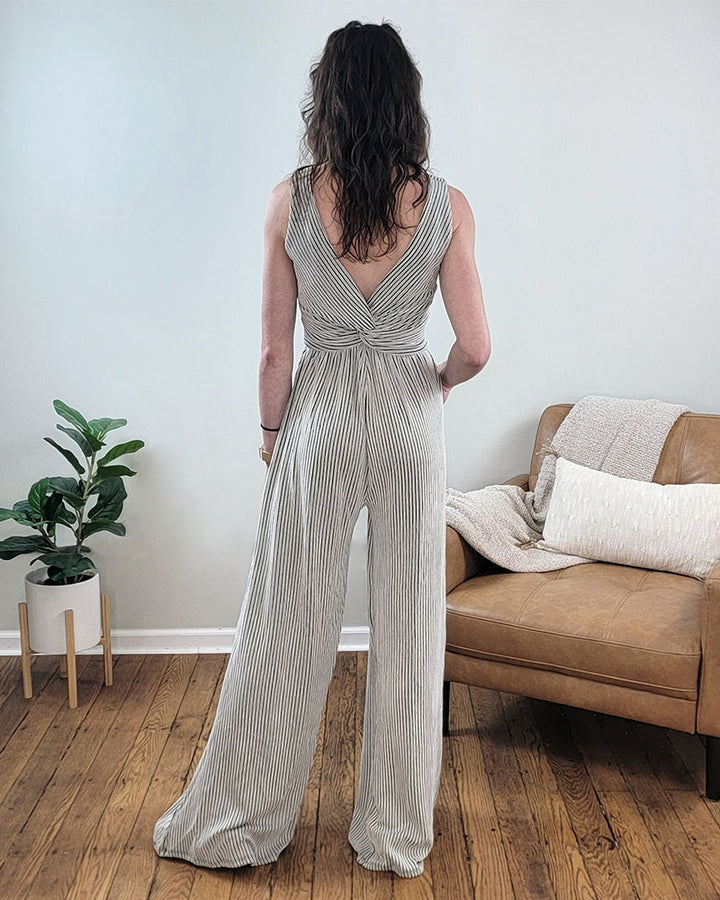 A woman with wavy brown hair, seen from behind, wears the Kaylee Romper by Selfmade Orange—a sleeveless, light gray-striped romper with a deep V-back—while standing on a wooden floor near a leather couch and potted plant.
