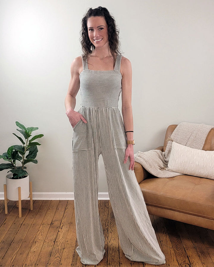 A woman stands indoors on a wooden floor, wearing the Selfmade Orange Kaylee Romper with wide legs and pockets. A potted plant and a tan couch with cushions and a throw blanket create a cozy backdrop.