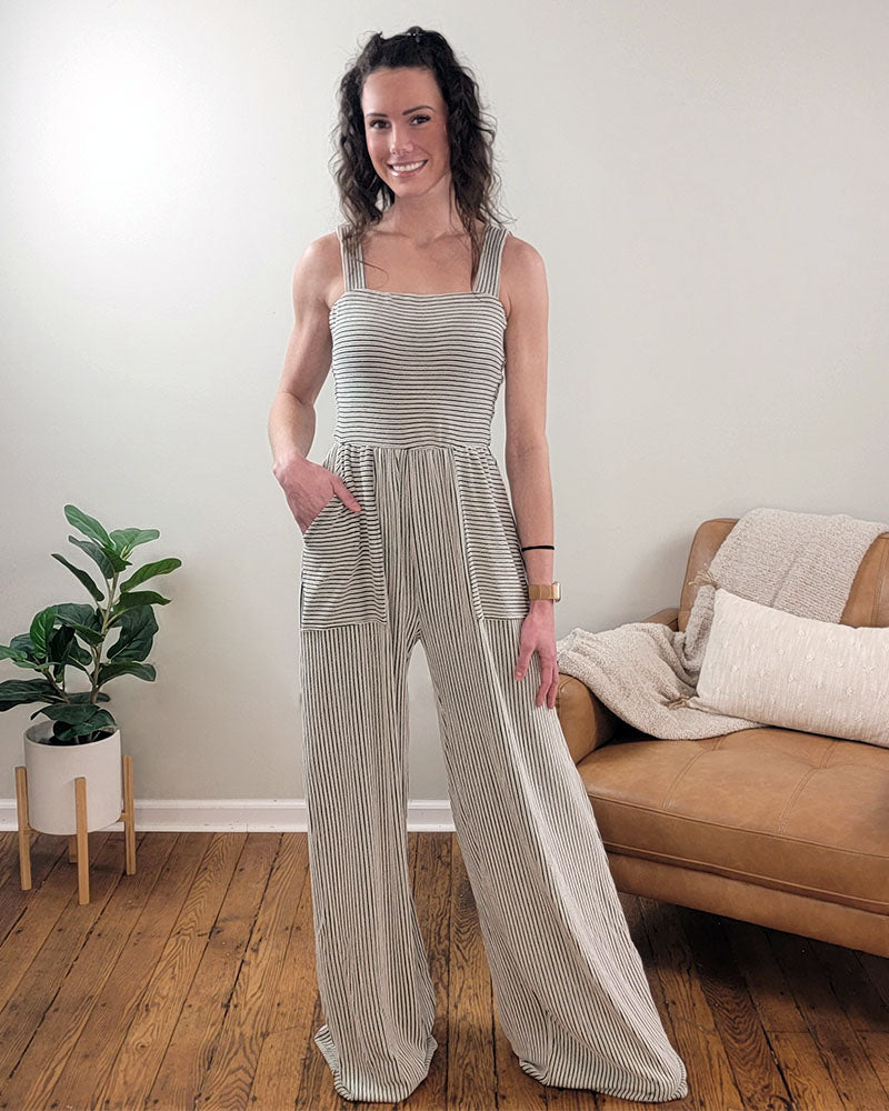 A woman stands indoors on a wooden floor, wearing the Selfmade Orange Kaylee Romper with wide legs and pockets. A potted plant and a tan couch with cushions and a throw blanket create a cozy backdrop.