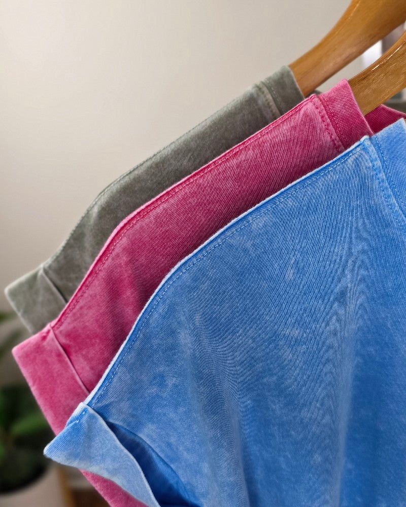 Three ZENANA Emma Snow Washed Short Sleeve Tops in blue, pink, and gray hang on wooden hangers, displayed side by side with a soft, blurred background. Each top features a relaxed fit design that blends comfort with effortless style.