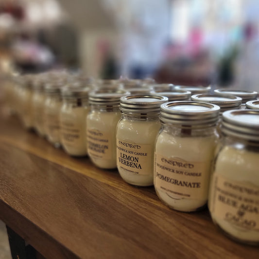 A row of Enspired Soy Candle jars with metal lids sits on a wooden shelf. Each handmade candle is labeled with scents like Lemon Verbena and Pomegranate, while the background remains tastefully blurred.