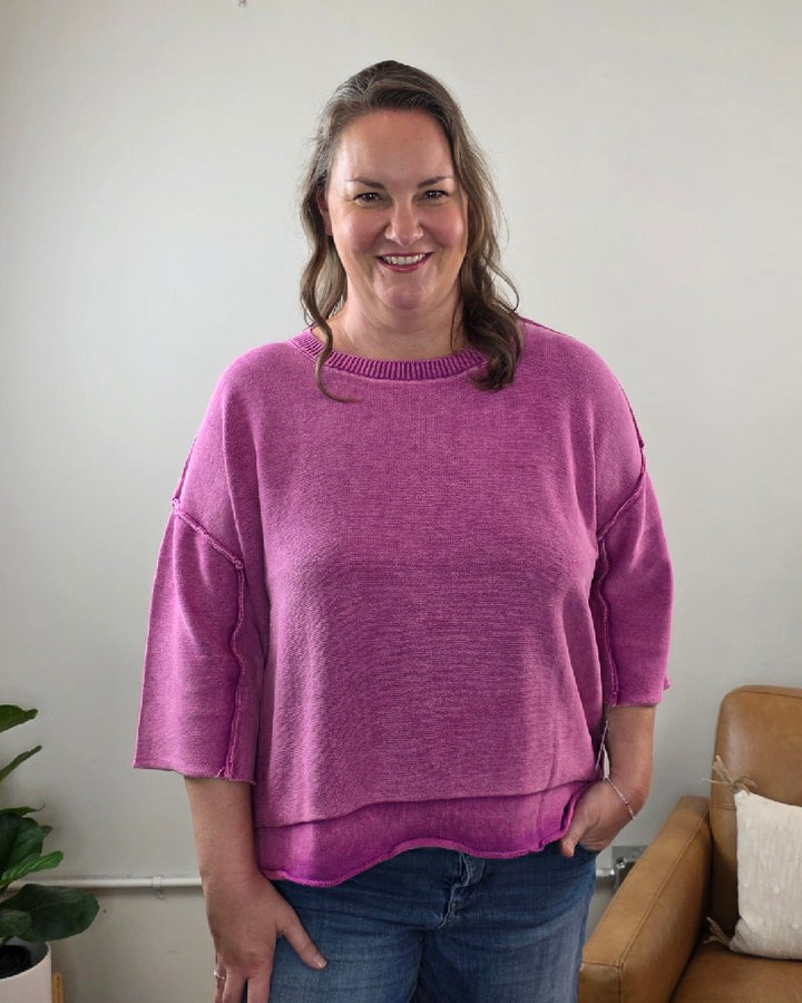A woman with wavy brown hair, dressed in the ZENANA Stacey 3/4 Sleeve Sweater With Exposed Seams in light plum and blue jeans, stands indoors smiling at the camera. A brown chair and a potted plant are visible in the background.