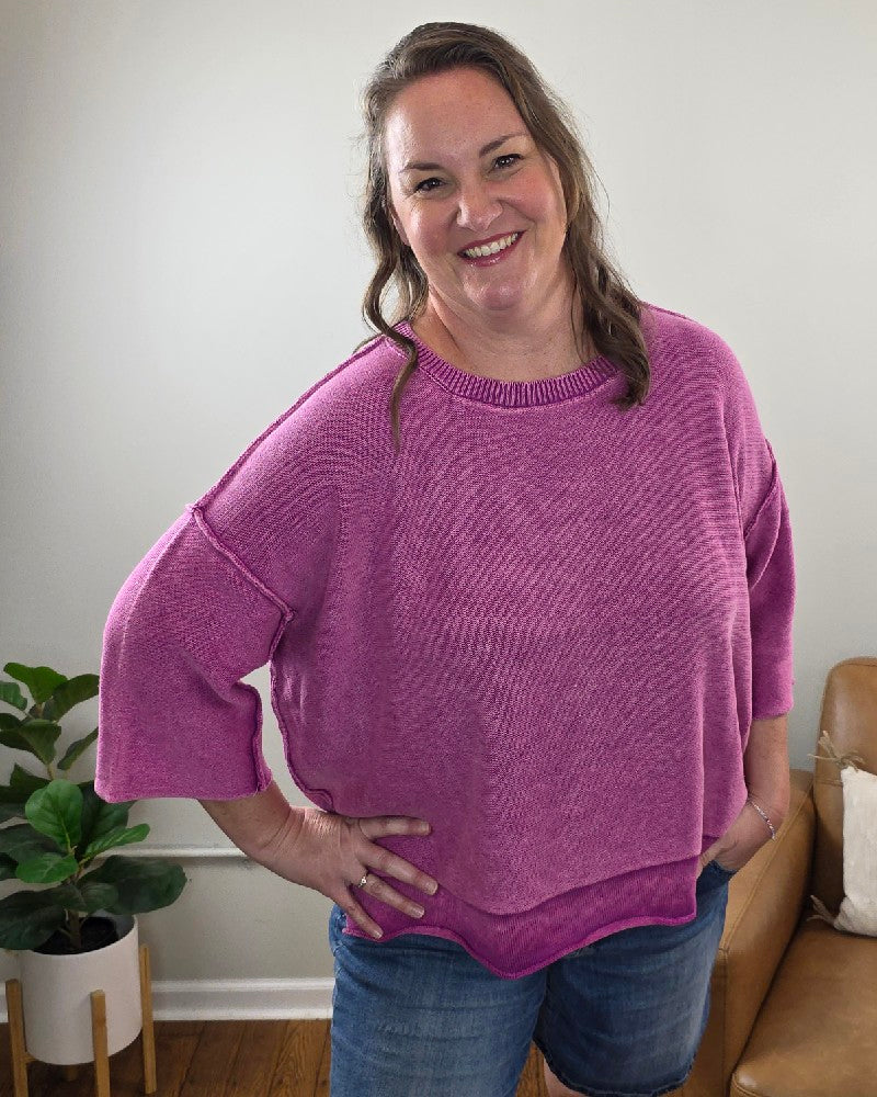 A woman with long brown hair is indoors, smiling with one hand on her hip, wearing the ZENANA Stacey 3/4 Sleeve Sweater With Exposed Seams in light plum and denim shorts. A potted plant and a brown armchair are visible in the background.