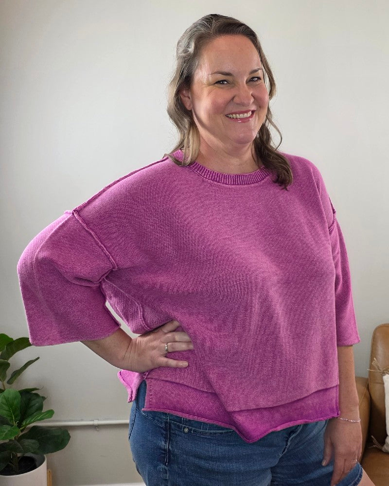 A woman with brown hair smiles indoors, wearing the ZENANA Stacey 3/4 Sleeve Sweater With Exposed Seams in a light plum shade and blue jeans. She poses with one hand on her hip, emphasizing the sweater’s exposed seam details. A green potted plant is visible in the background.