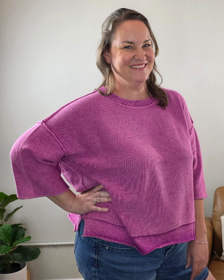 A woman with brown hair smiles indoors, wearing the ZENANA Stacey 3/4 Sleeve Sweater With Exposed Seams in a light plum shade and blue jeans. She poses with one hand on her hip, emphasizing the sweater’s exposed seam details. A green potted plant is visible in the background.