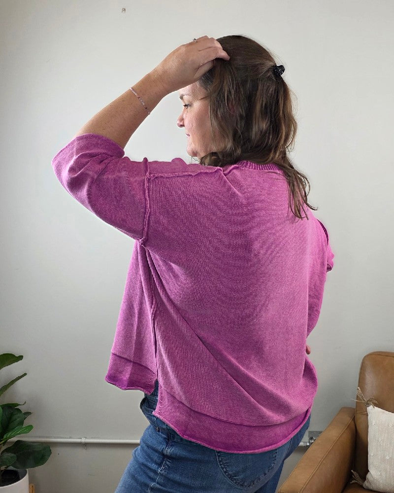 A woman with long brown hair styled half-up is seen from behind, wearing the ZENANA Stacey 3/4 Sleeve Sweater With Exposed Seams in light plum, paired with blue jeans. She stands with her left hand touching her head, near a tan chair and a green plant.