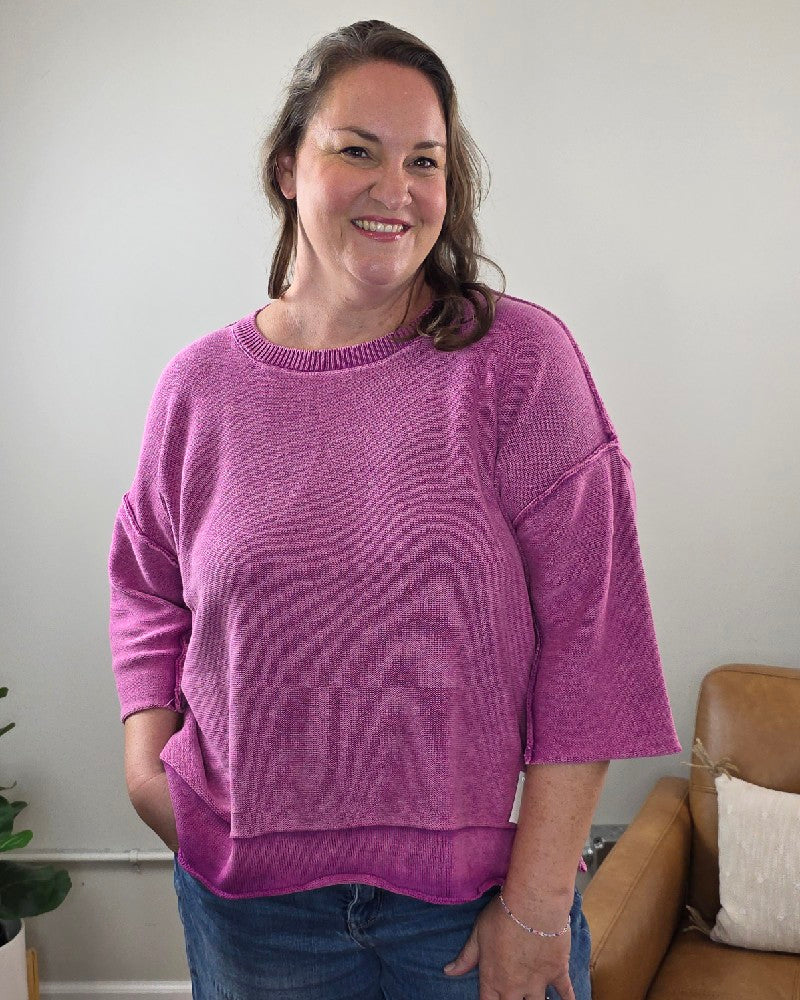 A woman with long brown hair smiles while standing indoors in a cozy living room with a tan chair and small plant in the background. She is wearing the ZENANA Stacey 3/4 Sleeve Sweater With Exposed Seams in light plum, paired with blue jeans.