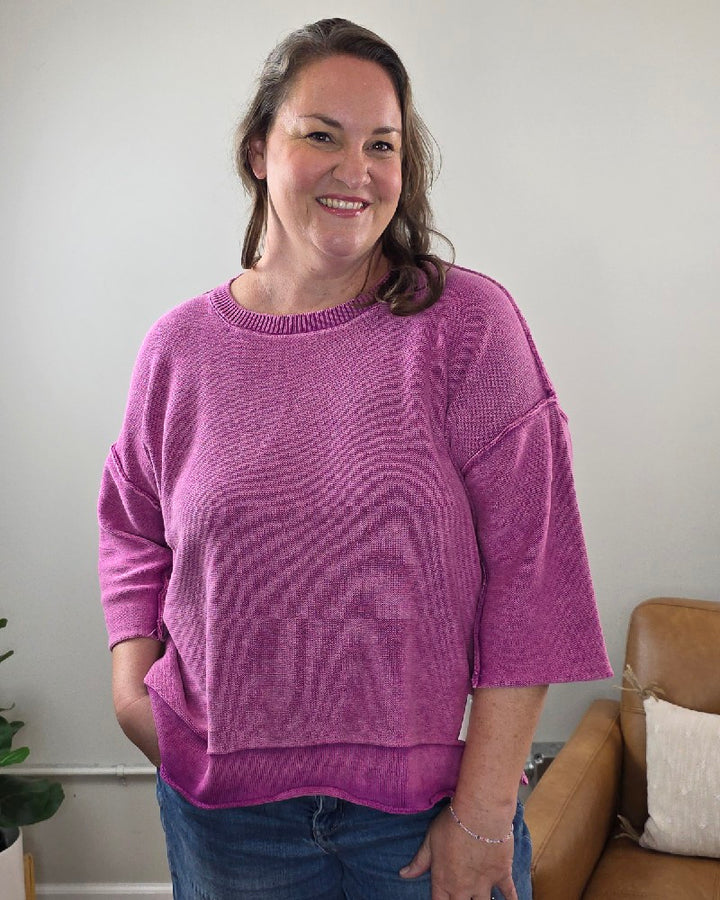 A woman with long brown hair smiles while standing indoors in a cozy living room with a tan chair and small plant in the background. She is wearing the ZENANA Stacey 3/4 Sleeve Sweater With Exposed Seams in light plum, paired with blue jeans.