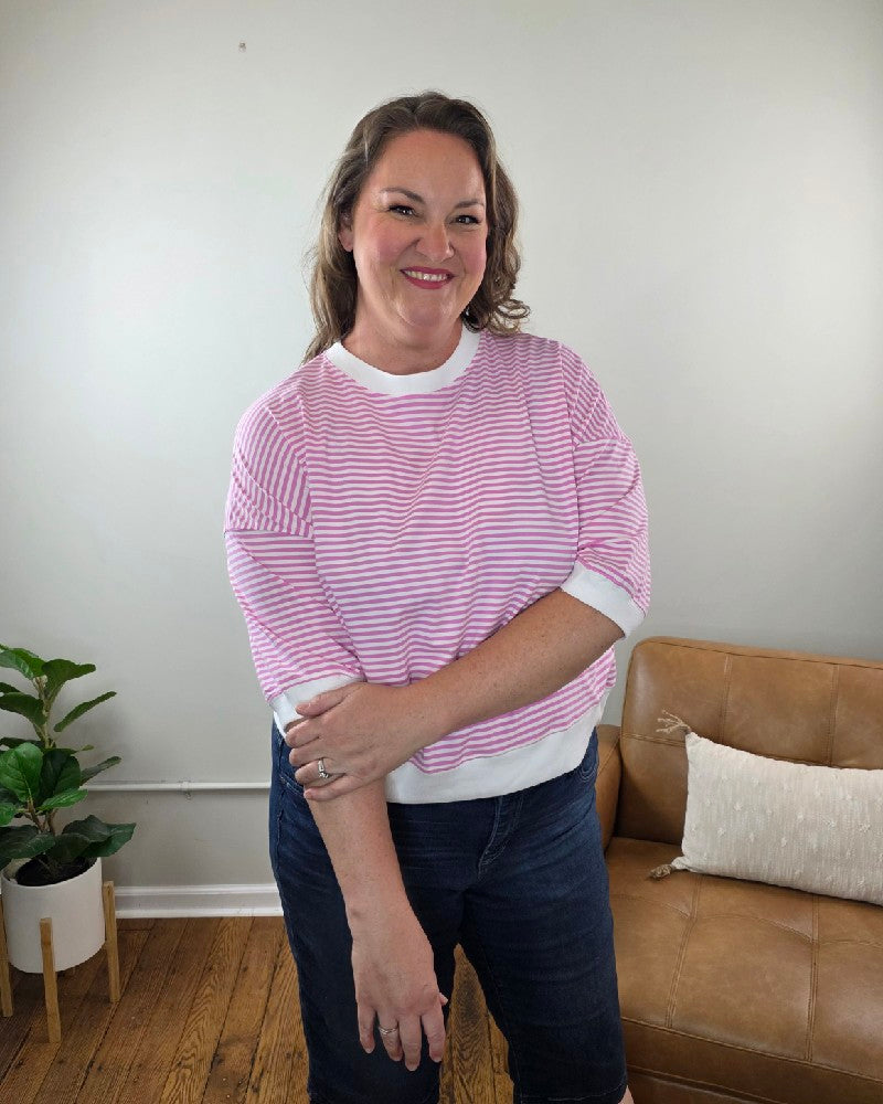 A woman with wavy brown hair smiles indoors, wearing the ZENANA Sylvia Striped Contrast Trim Rib Banded Top in candy pink and dark shorts. She stands by a tan couch with a light pillow and a green potted plant on the wooden floor.