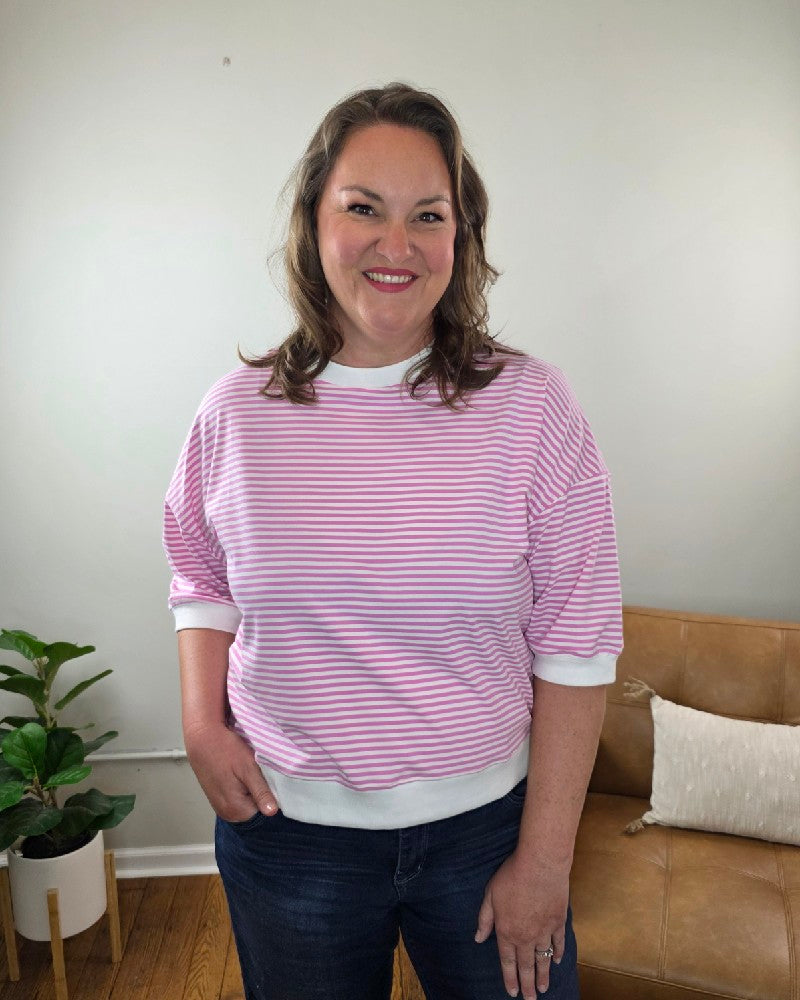 A woman with wavy brown hair smiles indoors, wearing the ZENANA Sylvia Striped Contrast Trim Rib Banded Top in candy pink paired with dark jeans. A tan couch with a pillow and a potted plant are visible in the background.