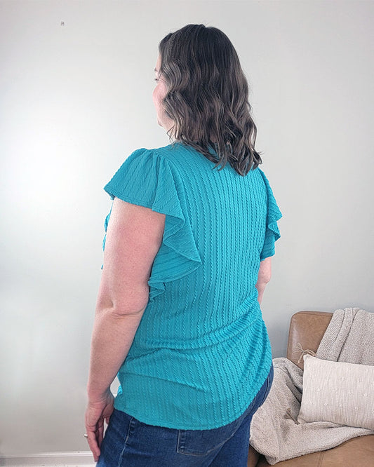 A woman with wavy brown hair stands indoors by a beige couch, wearing the Heimish Havanah Ruffle Sleeve Top in turquoise and blue jeans.