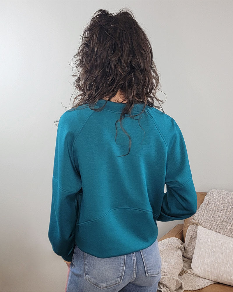 A person with wavy brown hair wearing the Yelete Group Maya Scuba Pullover in teal and blue jeans stands with their back to the camera in a cozy room with a beige couch and textured pillows.