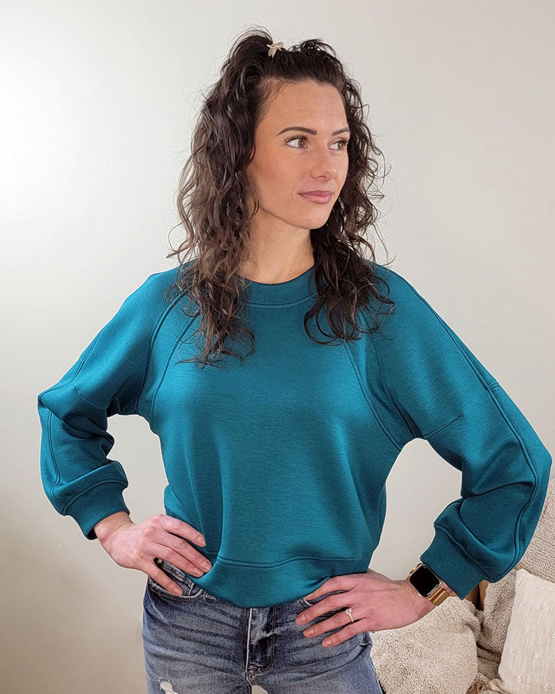 A woman with curly brown hair wears the Maya Scuba Pullover by Yelete Group—a cozy, cropped teal crewneck—paired with ripped jeans, standing against a light wall with her hands on her hips, looking to the side.