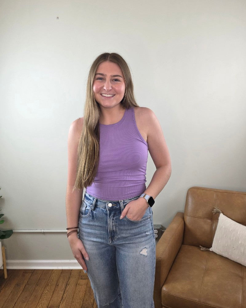 A young woman with long, straight hair smiles indoors, wearing the ZENANA Vicky Ribbed Racer Back Tank with Built In Bra in lavender and high-waisted jeans, one hand in her pocket. A tan couch is visible in the background.