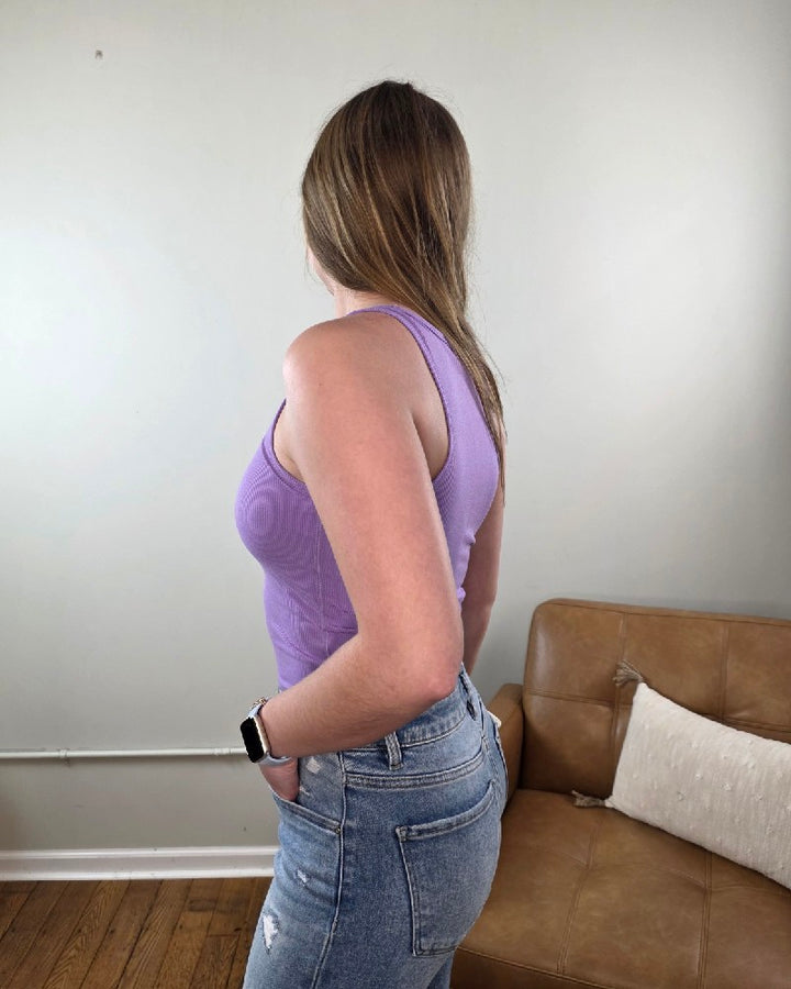 Woman wearing the Vicky ribbed, cropped, racerback tank top in lavender from Cosmos and Clover while standing in front of a tan chair. 