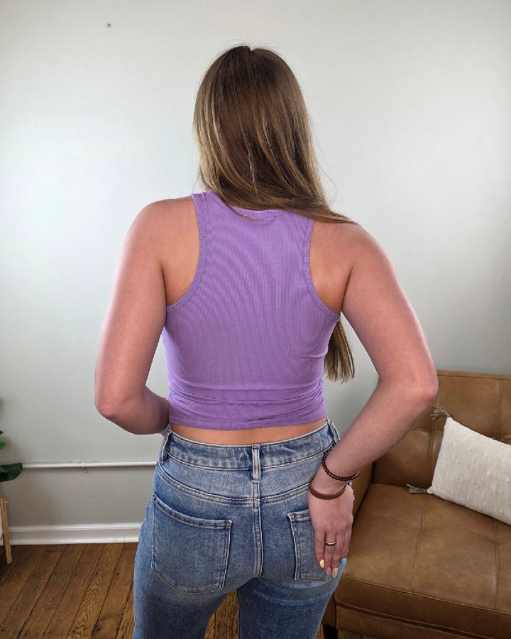 Woman wearing the Vicky ribbed, cropped, racerback tank top in lavender from Cosmos and Clover while standing in front of a tan chair. 