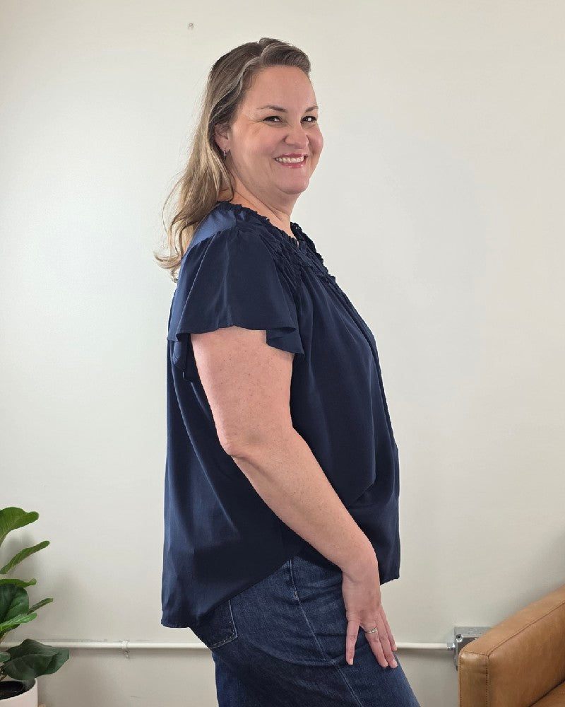 Woman with long hair stands sideways and smiles in a simple room, wearing the Vivian Detailed Blouse by blu pepper; a tan couch and green plant are visible in the background.