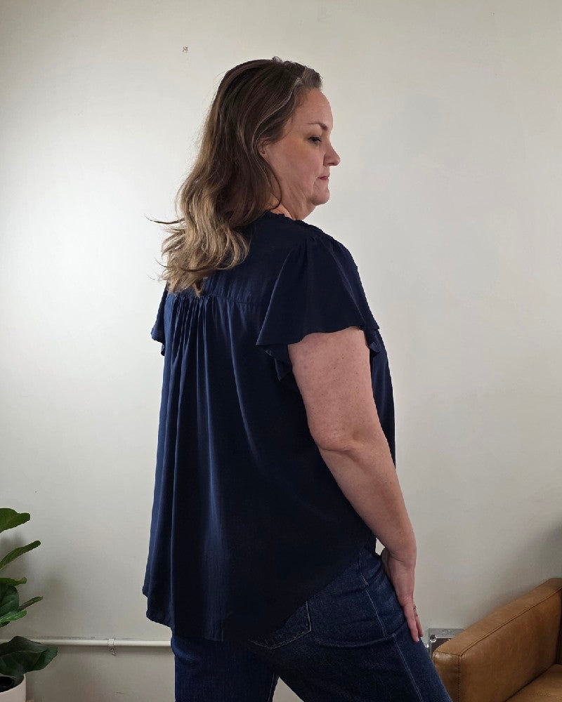 A woman with wavy brown hair stands sideways indoors, wearing the Vivian Detailed Blouse by blu pepper and blue jeans. A plant and part of a tan chair are visible against a neutral wall in the background.