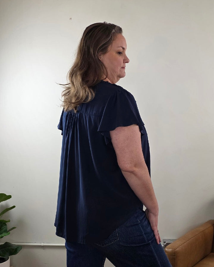 A woman with wavy brown hair stands sideways indoors, wearing the Vivian Detailed Blouse by blu pepper and blue jeans. A plant and part of a tan chair are visible against a neutral wall in the background.