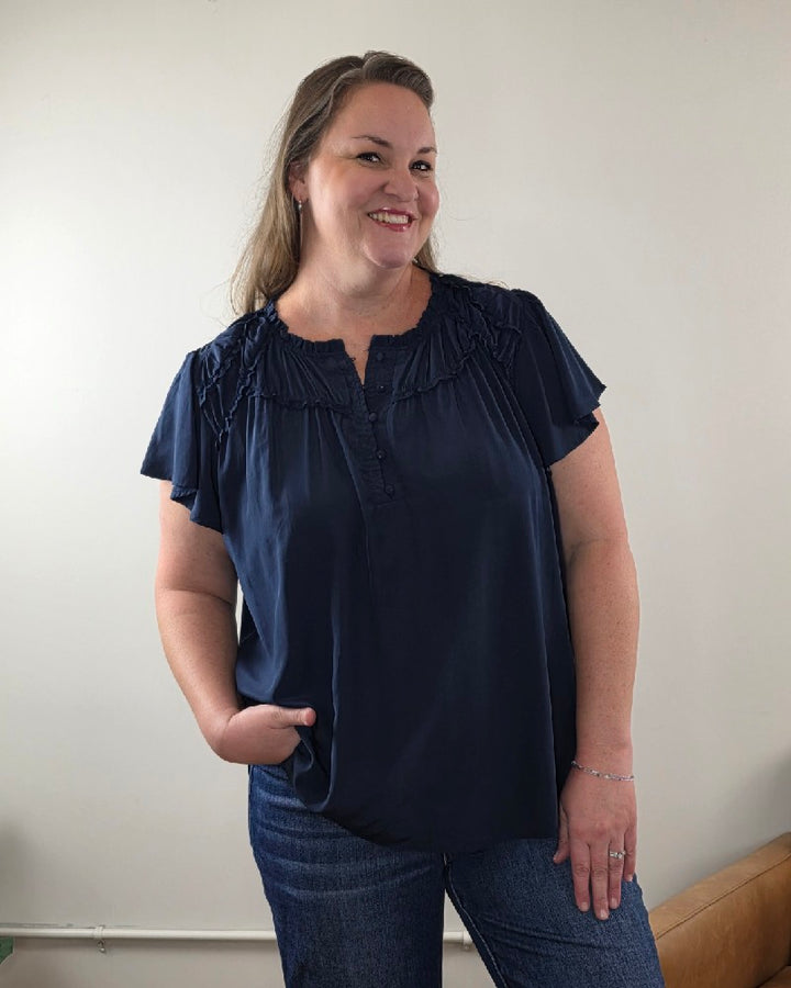 Woman with long hair smiles in the Vivian Detailed Blouse by blu pepper, paired with blue jeans, standing against a plain light-colored wall with one hand in her pocket.