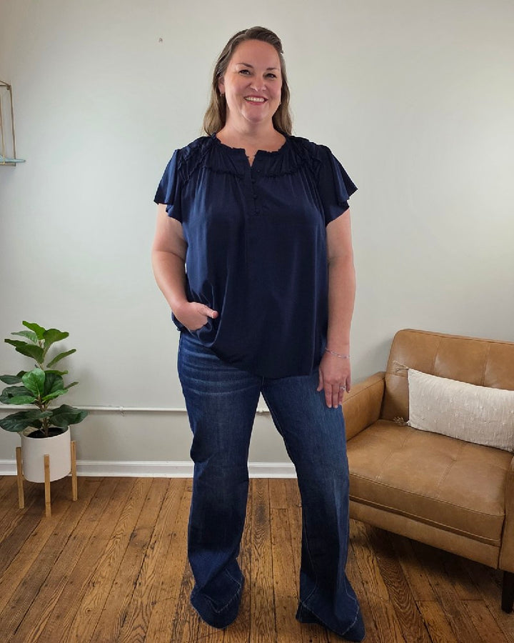 A woman smiles in a living room wearing the Vivian Detailed Blouse by blu pepper, paired with jeans. The room features wooden floors, a tan couch, and a potted plant.