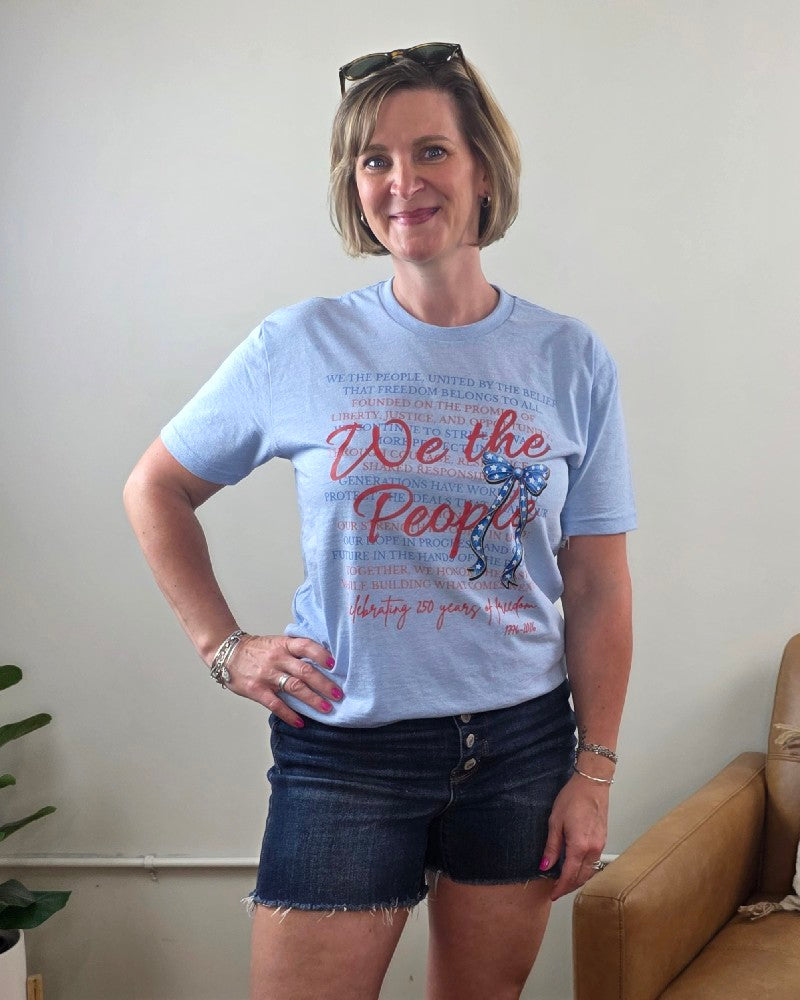 A smiling woman with short blonde hair wears the Cosmos + Clover Boutique "We The People" Graphic Tee and denim shorts, standing indoors beside a tan chair with one hand on her hip and sunglasses on her head.
