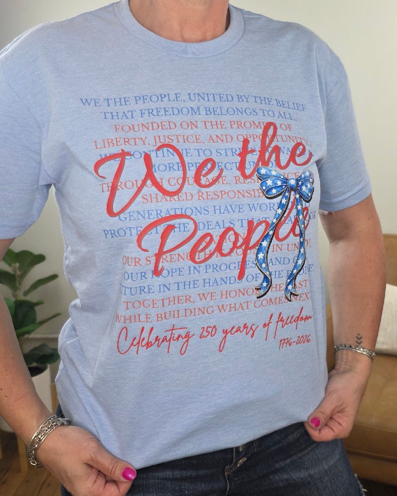 A person wears the Cosmos + Clover Boutique "We The People" Graphic Tee featuring bold red script, a blue star bow, and "Celebrating 250 years of freedom 1776-2026" in red at the bottom.