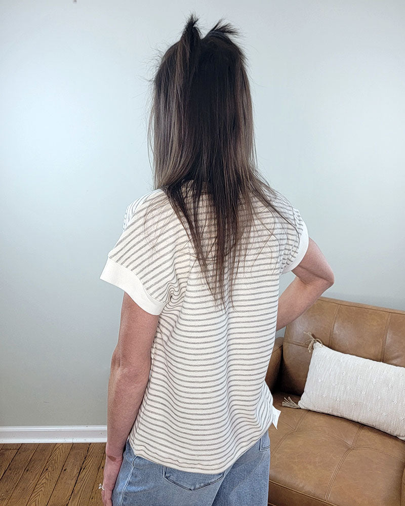 A person with long brown hair faces away, wearing the Staccato Posie Stripped Top—a 100% cotton, short-sleeved, light-striped shirt—and blue jeans. They stand near a tan couch with a white pillow in a room with light walls and wooden floors.