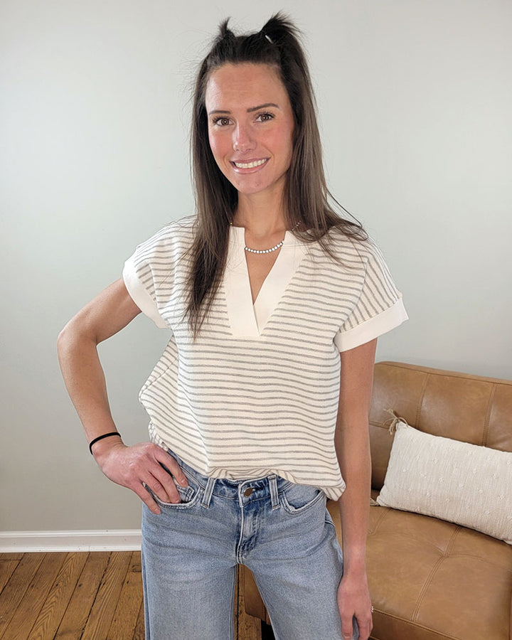 A woman with long brown hair smiles in a living room, wearing the Staccato Posie Striped Top—a short-sleeved, 100% cotton shirt—paired with light blue jeans. A tan couch and a pillow are visible in the background.
