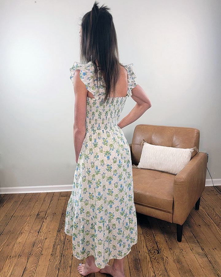 A woman with long brown hair stands barefoot on a wooden floor, wearing the Mine and E&M Lily Smocked Dress—featuring a light floral print, ruffled sleeves, and smocked bodice—in a simple room with a tan armchair and white pillow.