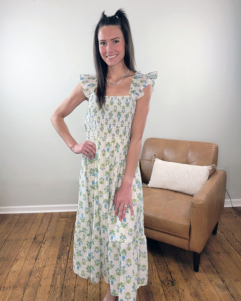 A woman stands on a wooden floor, smiling in the Mine and E&M Lily Smocked Dress—a white floral piece with blue and green patterns, ruffled sleeves, and a smocked bodice—posing next to a tan armchair with a white pillow.