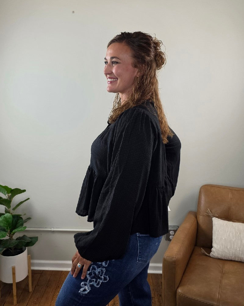 A woman with wavy brown hair smiles while standing sideways in a room with a potted plant, a leather chair, and white walls. She wears the HYFVE, Inc Willa Tie Front Peplum Blouse in black paired with blue jeans featuring floral embroidery.
