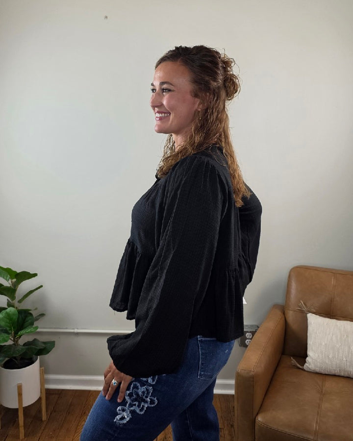 A woman with wavy brown hair smiles while standing sideways in a room with a potted plant, a leather chair, and white walls. She wears the HYFVE, Inc Willa Tie Front Peplum Blouse in black paired with blue jeans featuring floral embroidery.