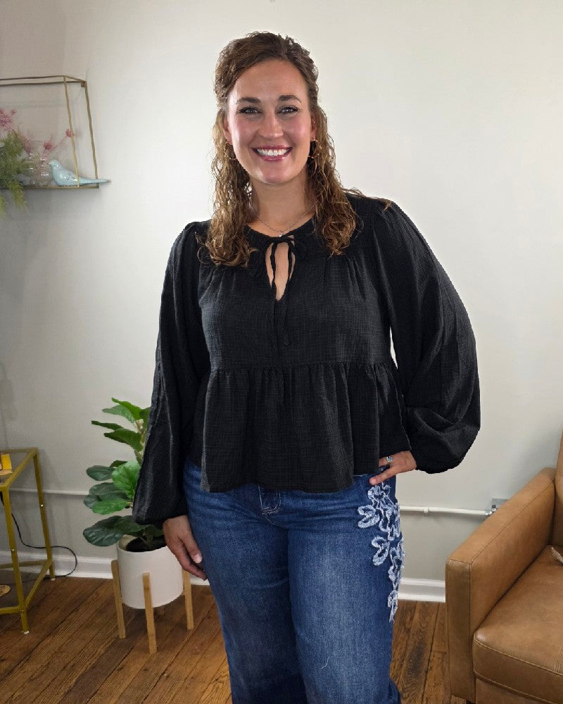 A woman with curly hair, dressed in the HYFVE, Inc Willa Tie Front Peplum Blouse in black and blue jeans adorned with white floral embroidery, stands smiling in a cozy room decorated with plants and a tan chair.
