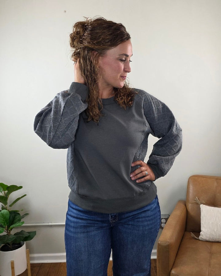 A woman with curly brown hair stands indoors, looking to the left with her hand touching her hair. She is wearing the Winona Contrast Sleeve Sweatshirt by Lime N Chili, Inc in a relaxed fit, paired with blue jeans. A plant and a tan chair are visible in the background.