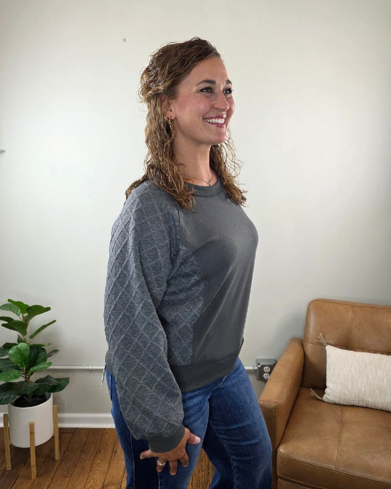 A woman with curly hair smiles while standing sideways in a room with a tan chair, pillow, and a potted plant. She wears the Winona Contrast Sleeve Sweatshirt by Lime N Chili, Inc—a relaxed fit, textured top in gray—paired with blue jeans.