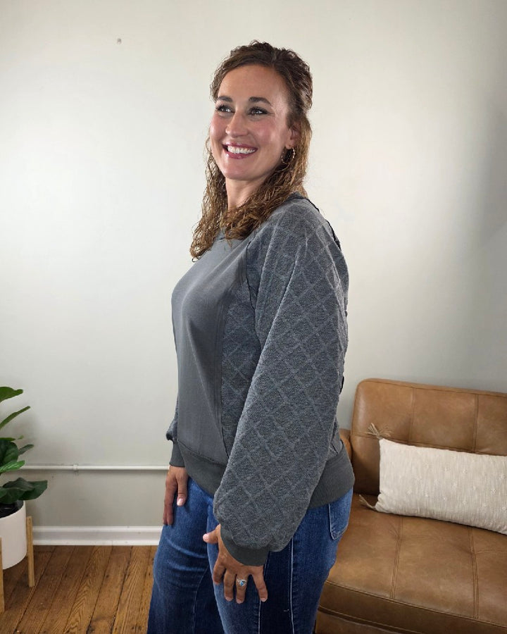A woman with wavy brown hair, wearing the Lime N Chili, Inc Winona Contrast Sleeve Sweatshirt and jeans, stands smiling in a room with a tan couch, white pillow, wooden floor, and a green potted plant.