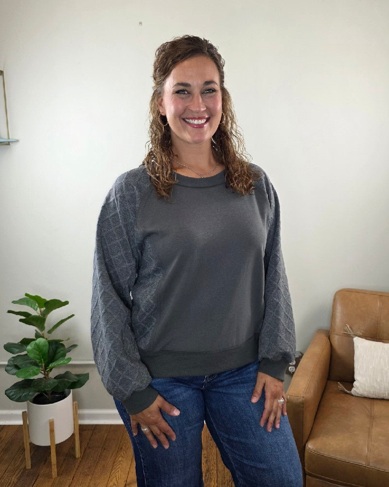A woman with curly hair stands smiling indoors on a wooden floor next to a brown chair and a potted plant, wearing the Winona Contrast Sleeve Sweatshirt by Lime N Chili, Inc with relaxed fit jeans.