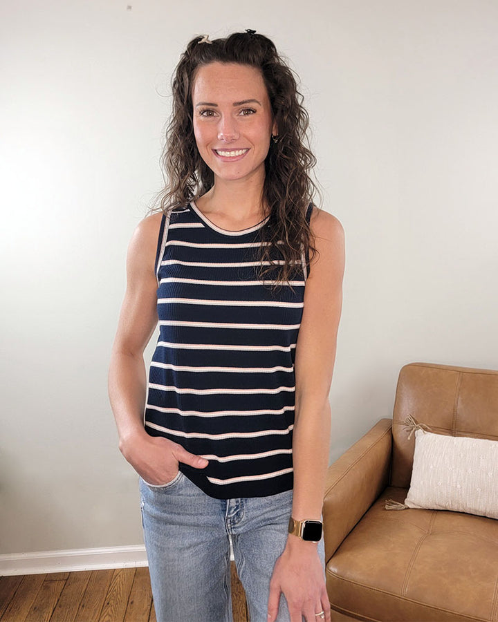 A woman with long curly hair smiles indoors, wearing the Staccato Carter Casual Striped Tank Top and jeans, with one hand in her pocket. A tan leather couch and pillow appear in the background.