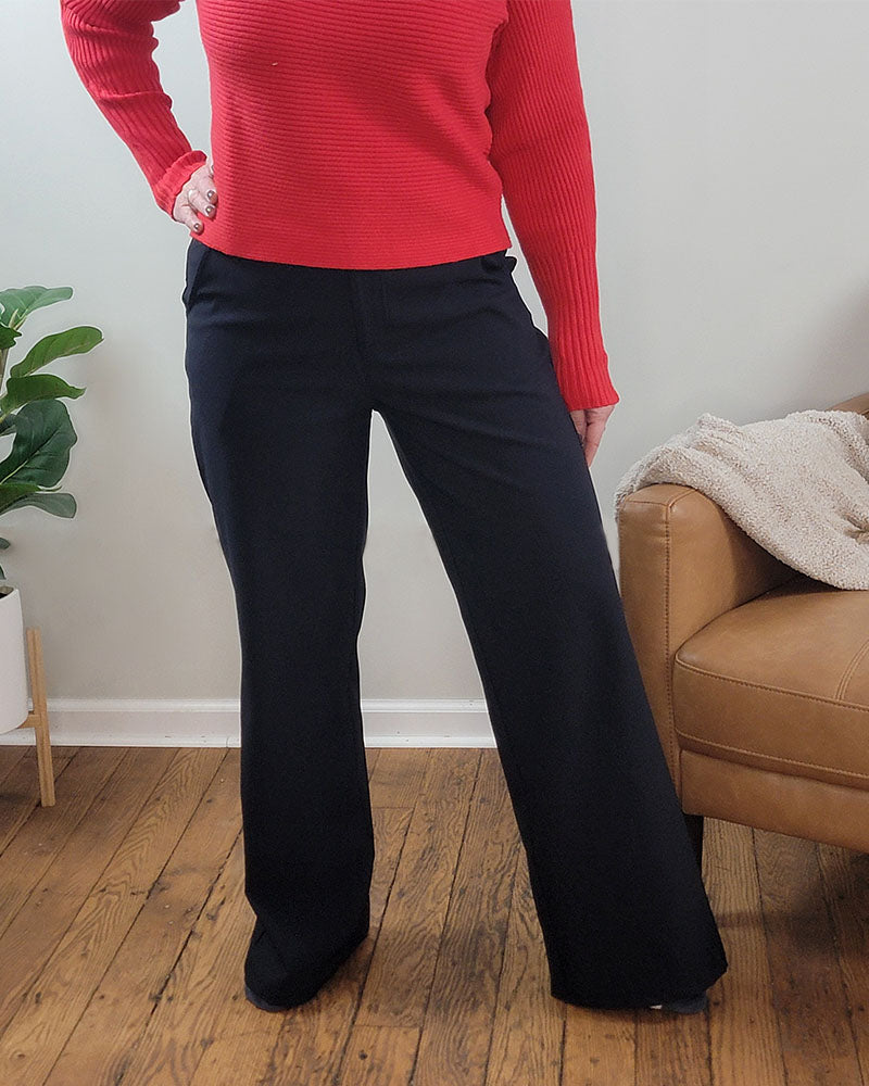 A person indoors stands on a wooden floor by a tan couch and green plant, wearing a red long-sleeve top and Mono B's Brenlee Wide Leg Pant in black.