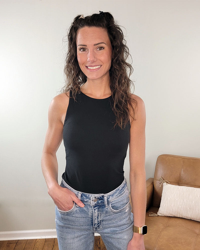 A woman with wavy brown hair wears the ZENANA Etta Bodysuit and blue jeans, smiling at the camera indoors. A tan couch with a white pillow appears in the background.