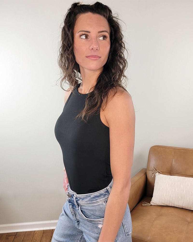 A woman with wavy brown hair wearing the ZENANA Etta Bodysuit in black and light blue jeans stands indoors beside a tan couch with a white pillow, looking to her left against a light-colored wall.
