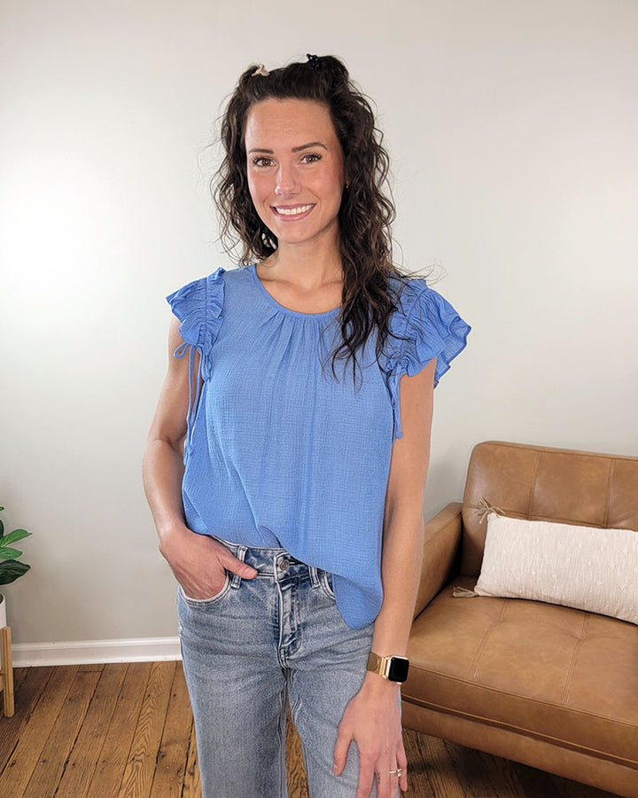 A woman with long wavy brown hair smiles indoors with one hand in her pocket, wearing the aqua Sadie Blouse by Mine and E&M and jeans. A tan couch and a potted plant are in the background.