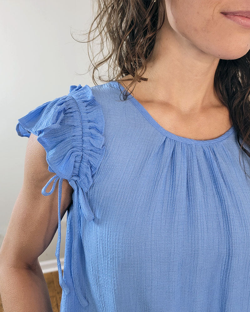 A woman stands indoors, partially out of frame, wearing the Sadie Blouse by Mine and E&M—an aqua top with a round neckline, ruffled shoulders, and gathered fabric.