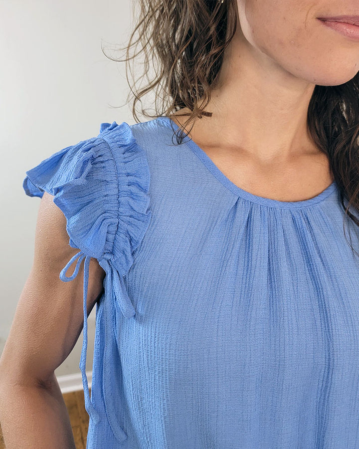 A woman stands indoors, partially out of frame, wearing the Sadie Blouse by Mine and E&M—an aqua top with a round neckline, ruffled shoulders, and gathered fabric.