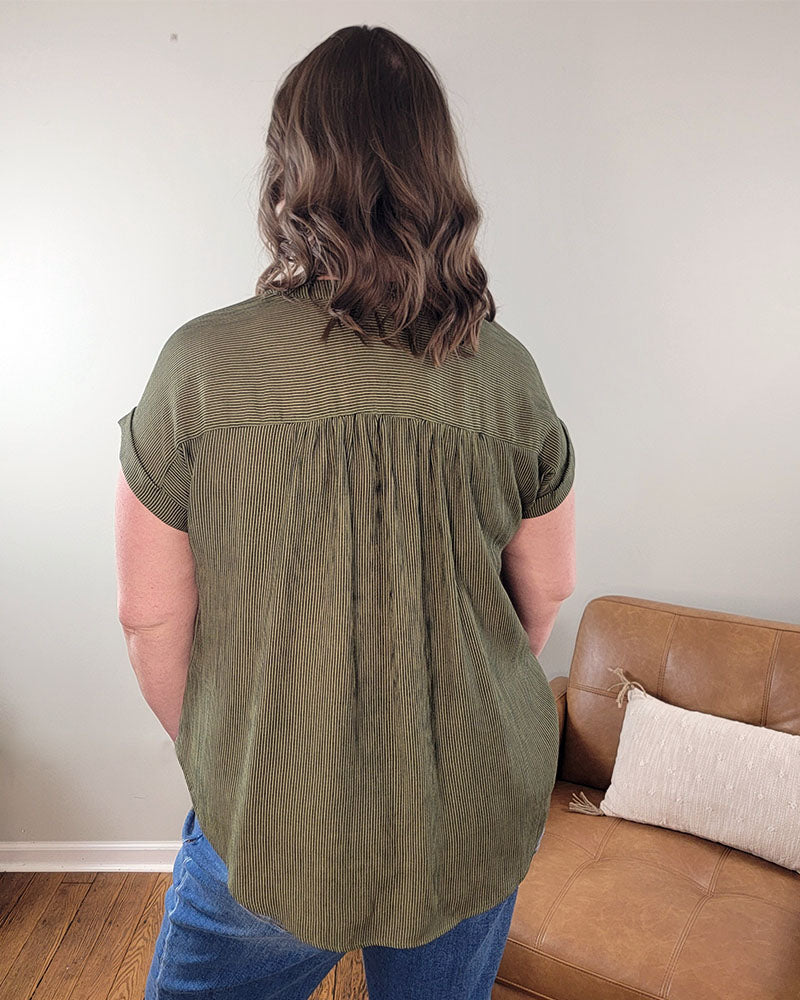 A person with wavy brown hair stands facing away from the camera, wearing the Emerson Button Front Top by Mine and E&M in olive green—short-sleeved and perfect for spring and summer—near a tan sofa with a white pillow.