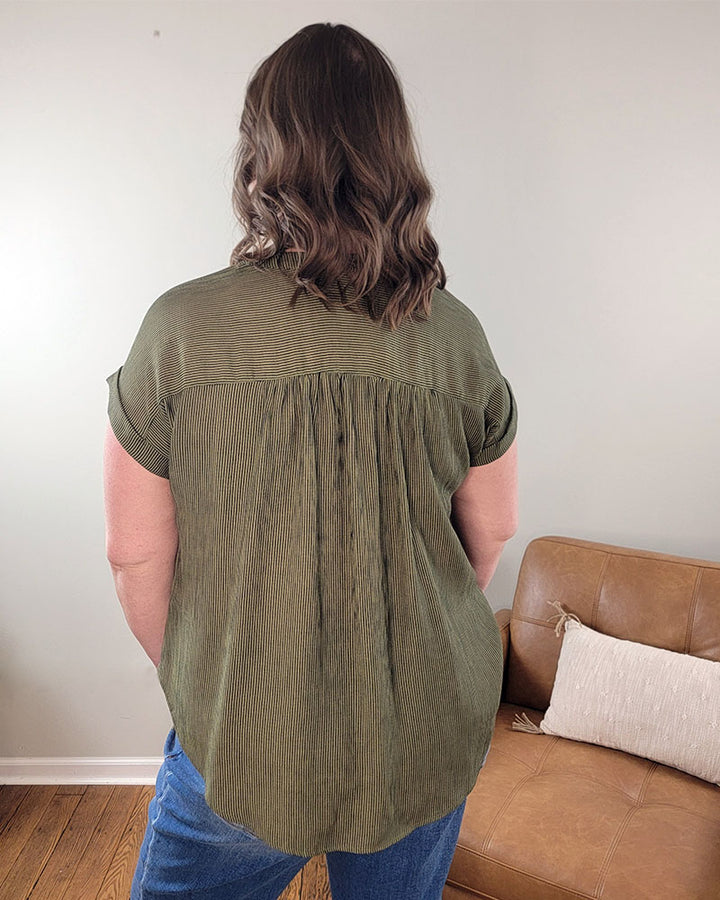 A person with wavy brown hair stands facing away from the camera, wearing the Emerson Button Front Top by Mine and E&M in olive green—short-sleeved and perfect for spring and summer—near a tan sofa with a white pillow.