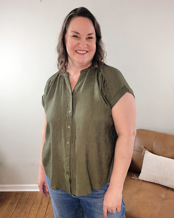 A woman with wavy brown hair smiles indoors, wearing the Mine and E&M Emerson Button Front Top in olive green with blue jeans, standing by a tan couch with a cream pillow—an ideal addition to your spring and summer wardrobe.
