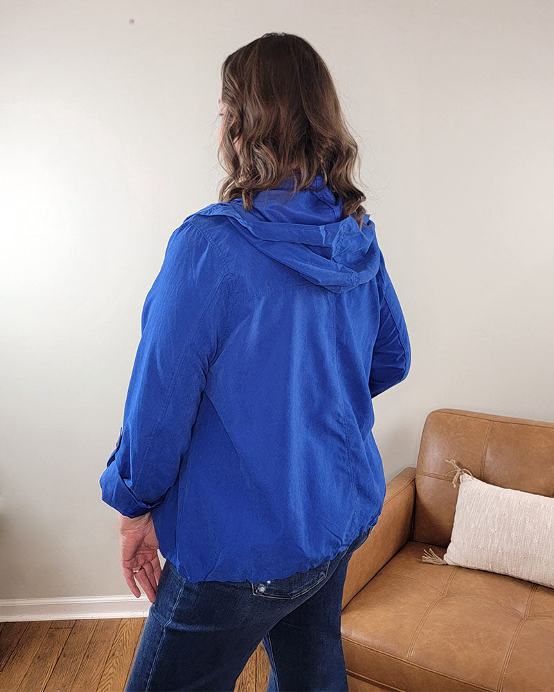 A person with wavy brown hair, wearing Mine and E&M's bright blue Alyson Jacket and jeans, stands indoors facing away from the camera next to a tan leather sofa with a beige pillow.